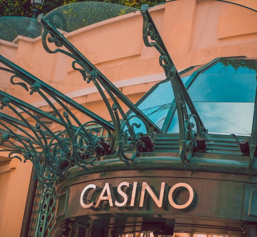 The image displays the entrance to a casino, characterized by detailed and ornate architectural elements with green metal structures and a sign that reads 'CASINO'. The background includes a glass dome and a peach-colored wall.