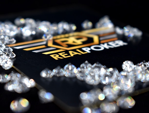 A collection of sparkling, clear gemstones surrounds a dark surface featuring the prominent logo and text 'Real Poker.' The logo design includes gold and white elements set against a black background.