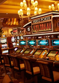 A casino floor featuring rows of brightly lit video poker machines. The area is illuminated by elegant chandeliers, with a display board showing a large sum of money, suggesting a possible jackpot. The rich decor includes luxurious carpets and comfortable seating, adding to the opulent atmosphere.