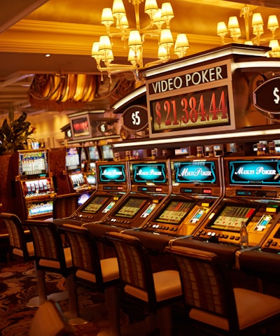 A casino floor featuring rows of brightly lit video poker machines. The area is illuminated by elegant chandeliers, with a display board showing a large sum of money, suggesting a possible jackpot. The rich decor includes luxurious carpets and comfortable seating, adding to the opulent atmosphere.