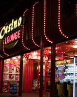A brightly lit casino lounge entrance adorned with neon lights. The exterior features colorful signage including the words 'Casino' and 'Lounge' in vivid colors, with additional lights framing the building. Inside, a glimpse of a claw machine filled with plush toys and arcade games is visible. The area exudes an inviting yet bustling atmosphere typical of entertainment venues.