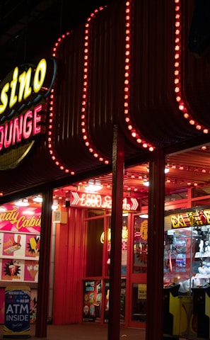 A brightly lit casino lounge entrance adorned with neon lights. The exterior features colorful signage including the words 'Casino' and 'Lounge' in vivid colors, with additional lights framing the building. Inside, a glimpse of a claw machine filled with plush toys and arcade games is visible. The area exudes an inviting yet bustling atmosphere typical of entertainment venues.