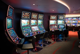 A row of brightly lit slot machines in a casino setting, each with colorful displays and various game themes. The machines are lined up against a dark wall and are unoccupied, with cushioned chairs placed in front of them. The area is dimly lit except for the glow from the screens, creating a vibrant and engaging atmosphere.