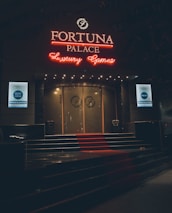 A neon-lit casino entrance with a red carpet leading up to large double doors. The sign reads 'Fortuna Palace Luxury Games' in bright red and white neon lights. The dimly lit atmosphere suggests a night-time setting. There are two digital displays on either side of the entrance showing promotional content.