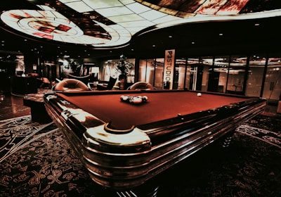 A luxurious interior features a billiard table with balls arranged on it. The ceiling has an intricate stained glass design, and the space is dimly lit, creating a sophisticated atmosphere. Comfortable chairs are placed around the area, with some decorative plants and a lamp adding to the elegance.
